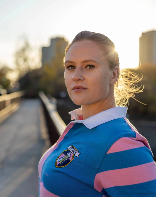 A blond woman wearing a pink and blue long sleeve rugby shirt with sociable crest