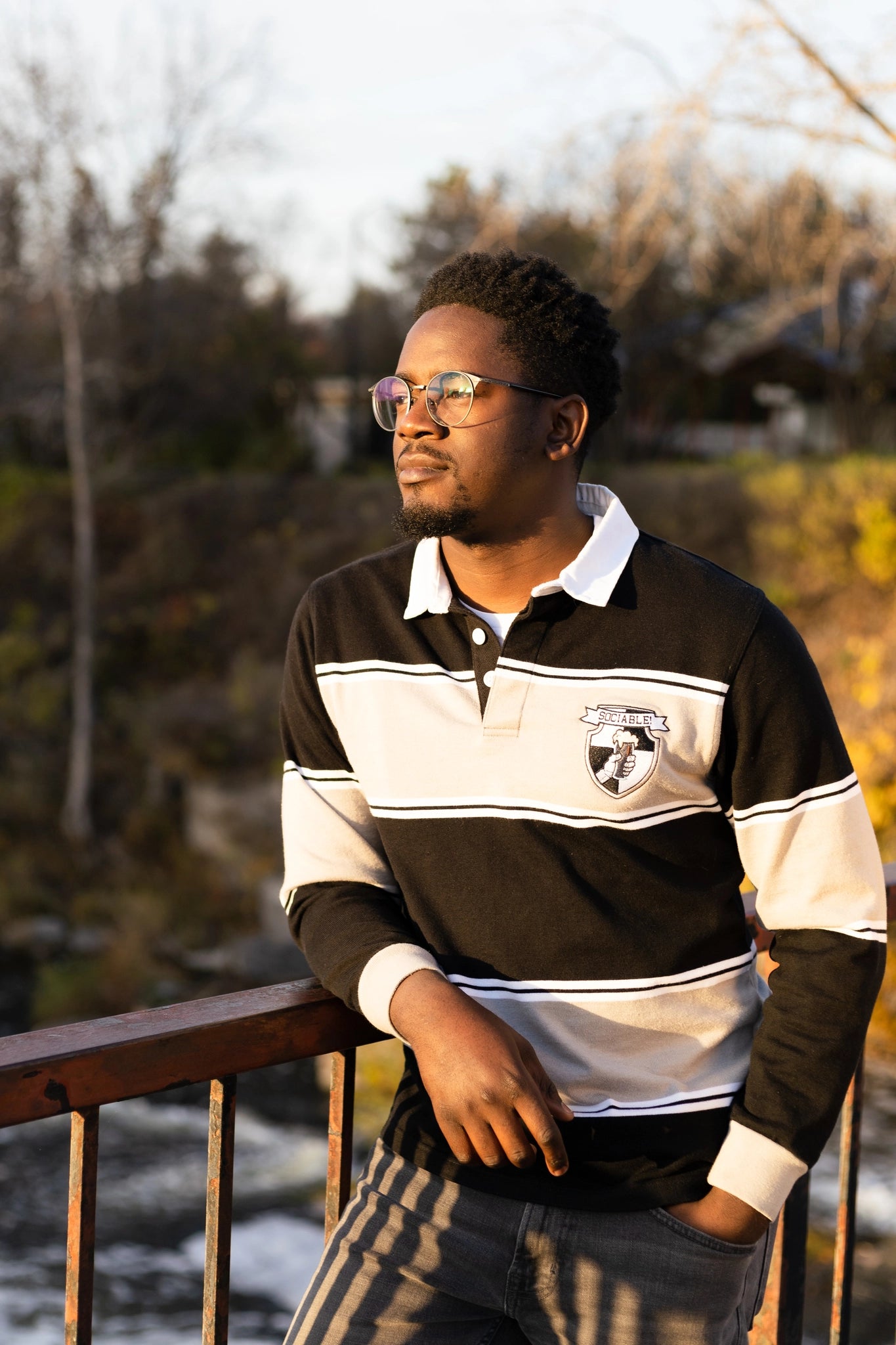 A black man wearing glasses on a bridge wearing a black and grey sociable rugby shirt